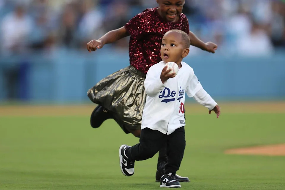 Kaj Betts wearing a Dodgers jersey runs away with a baseball, with big sister Kynlee wearing a sparkly dress close behind