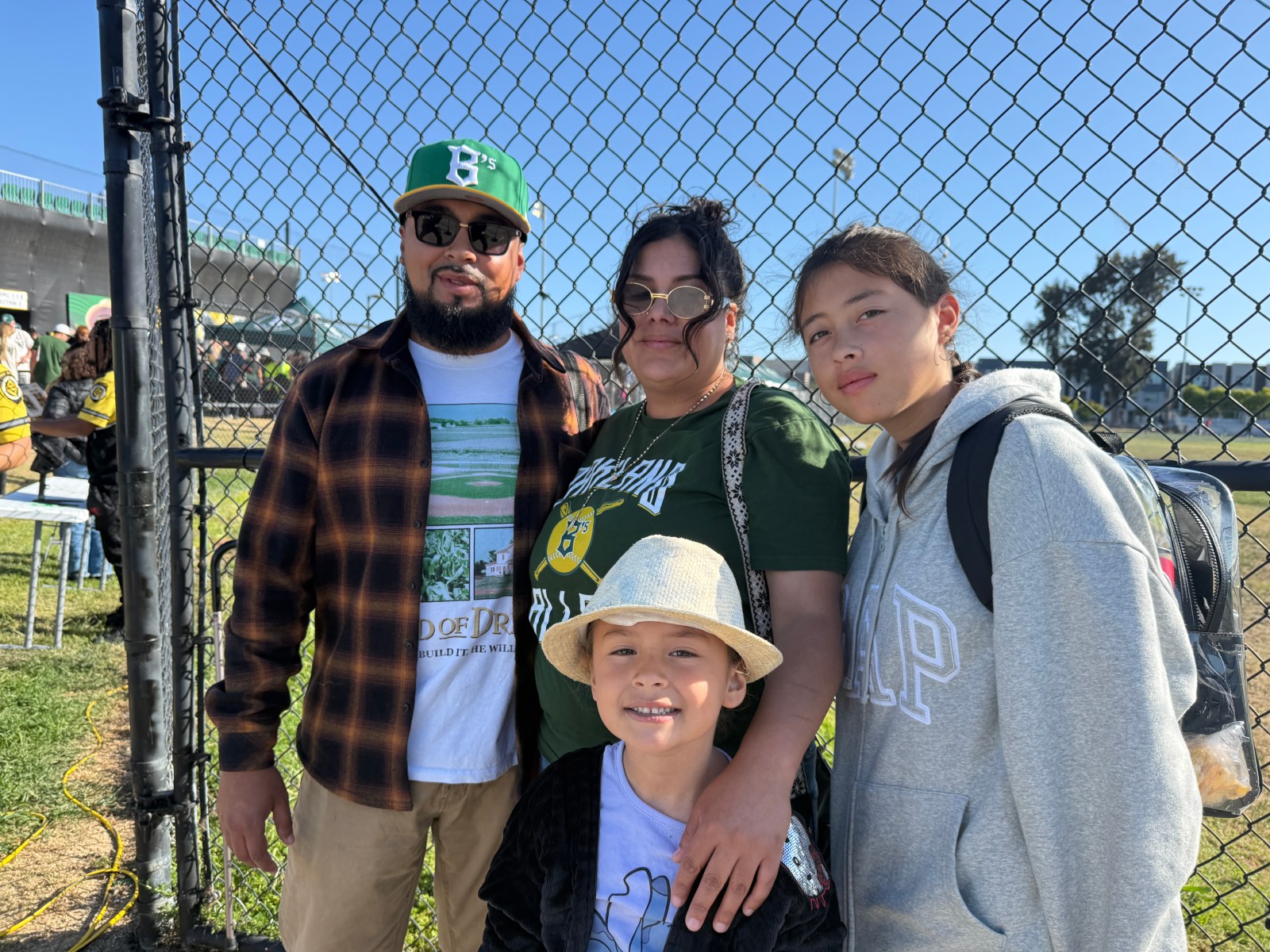 A family of four posing for a photo near a chain-link fence at a baseball field, smiling and wearing casual clothing, with sunny weather in the background.