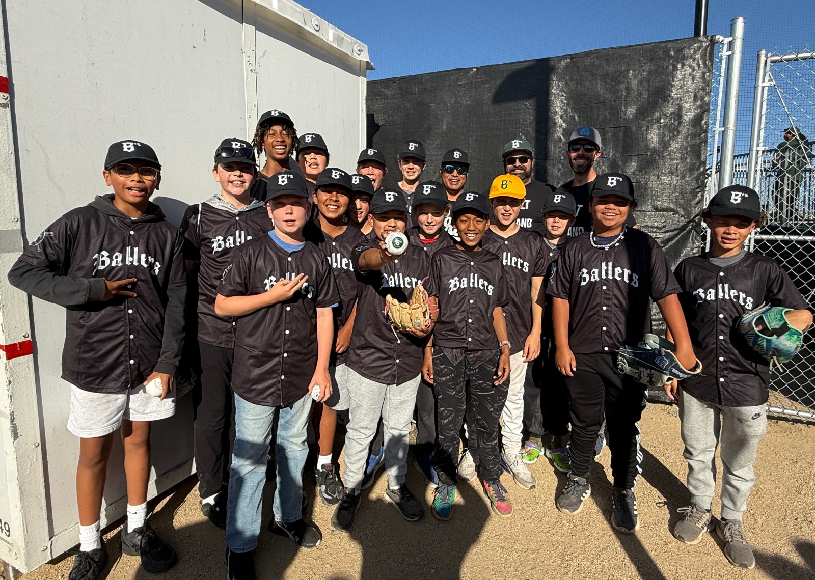 A group of young baseball players in matching black jerseys and caps, smiling and posing for a photo together, with a a glove and baseball in hand, and a sporting background.
