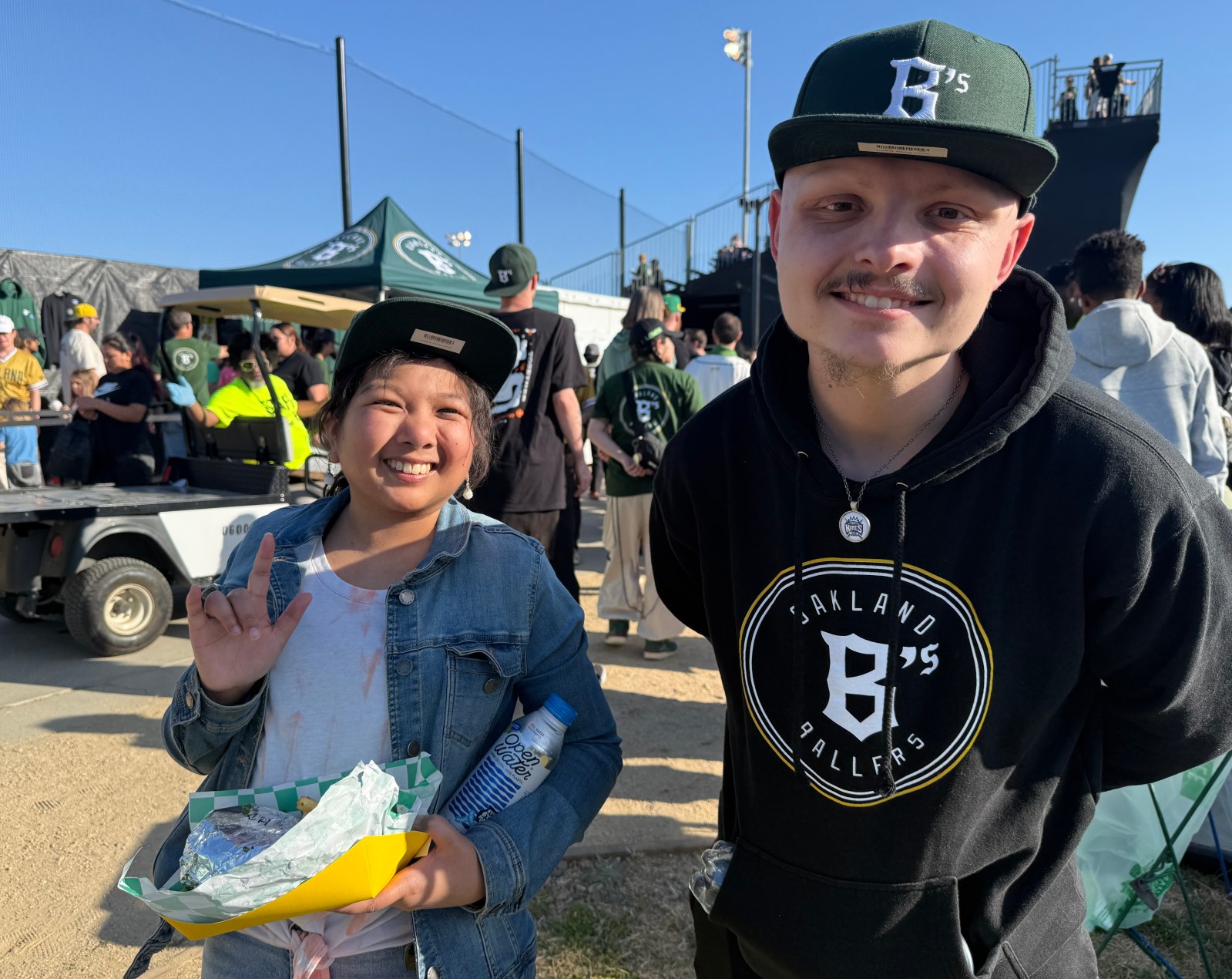 Two smiling fans of the Oakland Ballers pose for a photo, one holding a food tray and a water bottle, at a vibrant outdoor event.