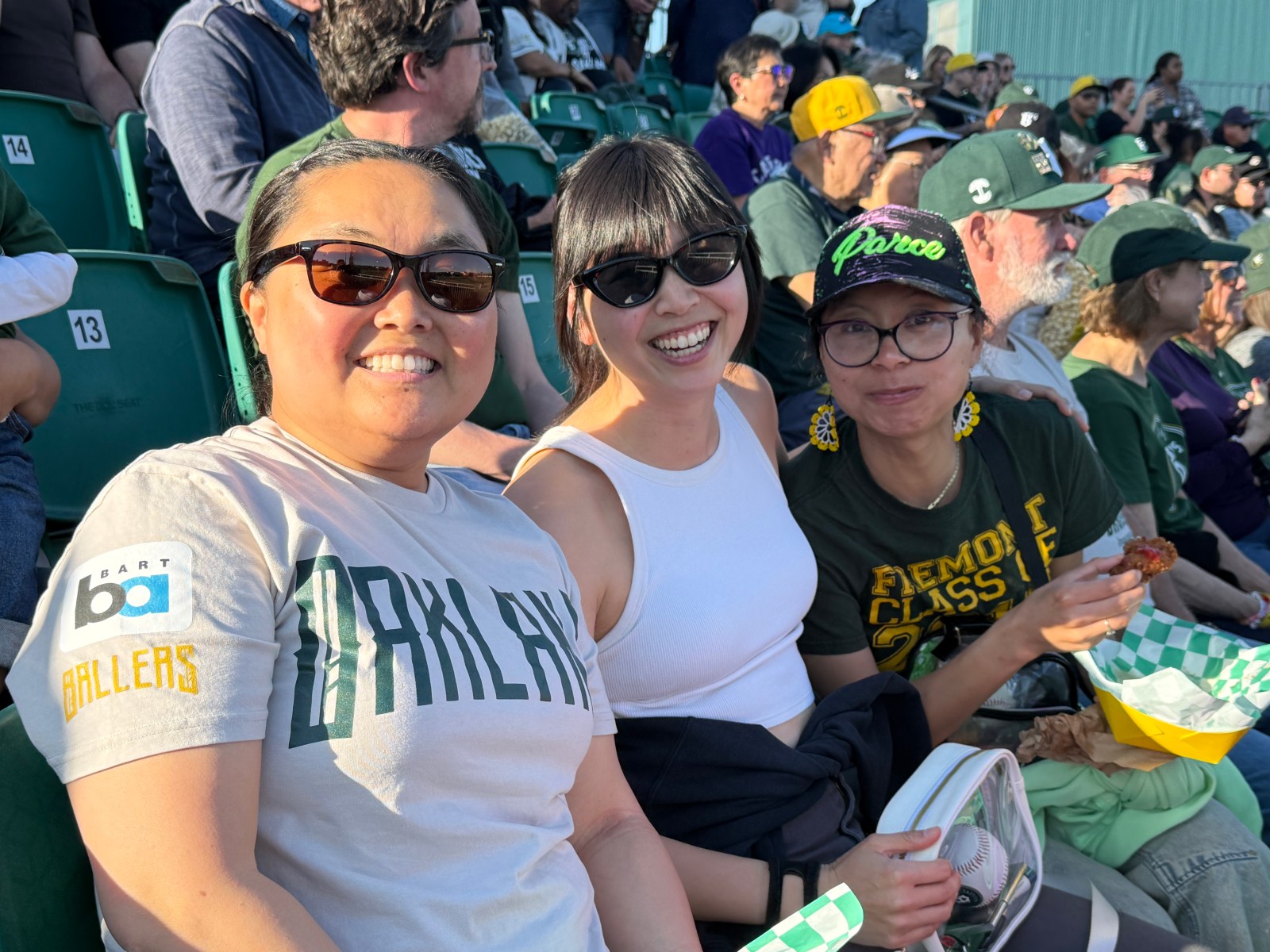 Three fans enjoying a baseball game at Raimondi Field, wearing Oakland Ballers clothing. The fan on the left is in a gray shirt with 'Oakland Ballers' printed, while the middle fan is wearing a white top. The fan on the right is wearing dark green and holding a piece of food. They are smiling, amidst a crowd.