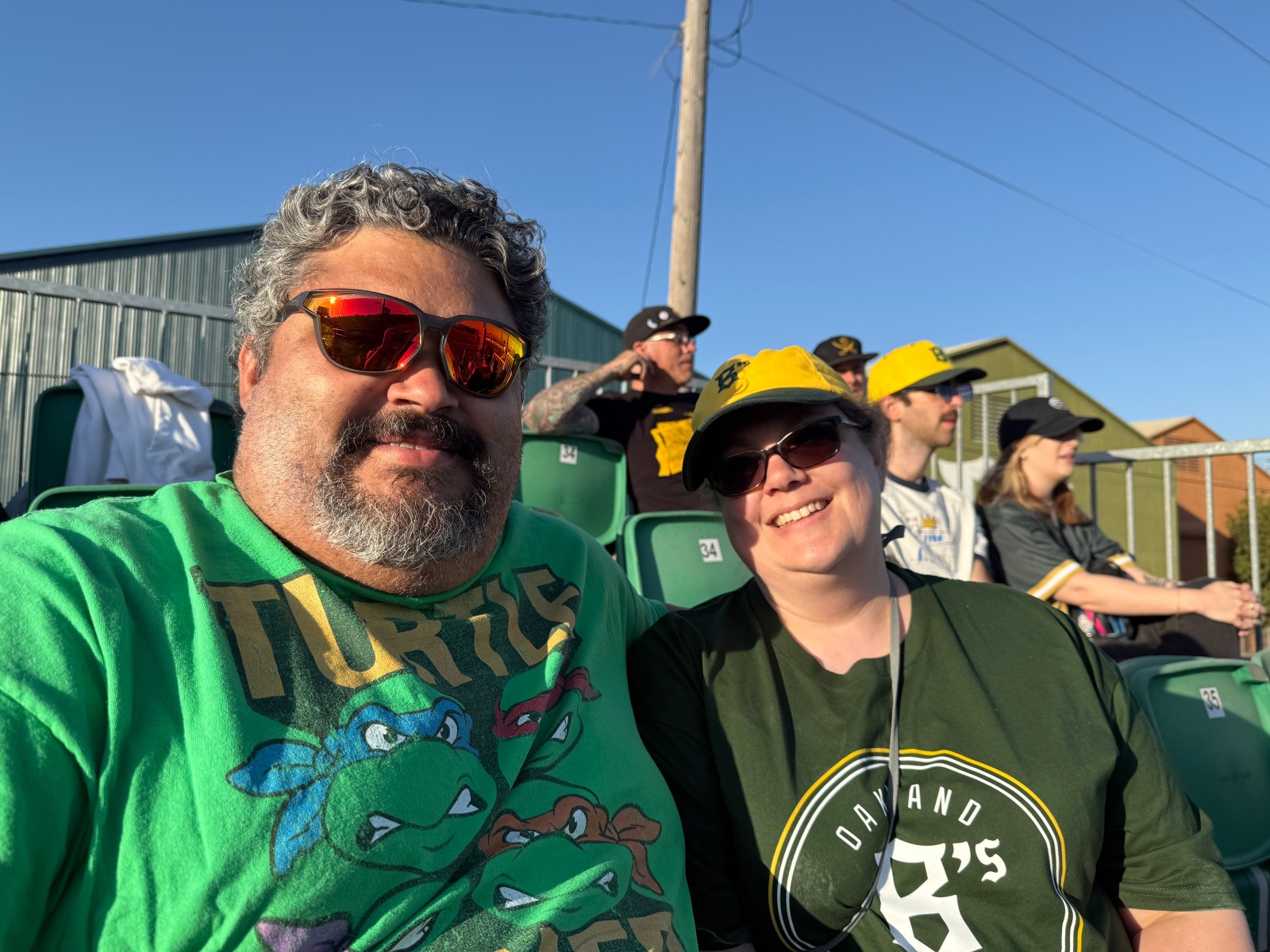 A man and a woman pose for a photo in the stands at a baseball game, wearing team shirts and sunglasses, with other fans visible in the background.