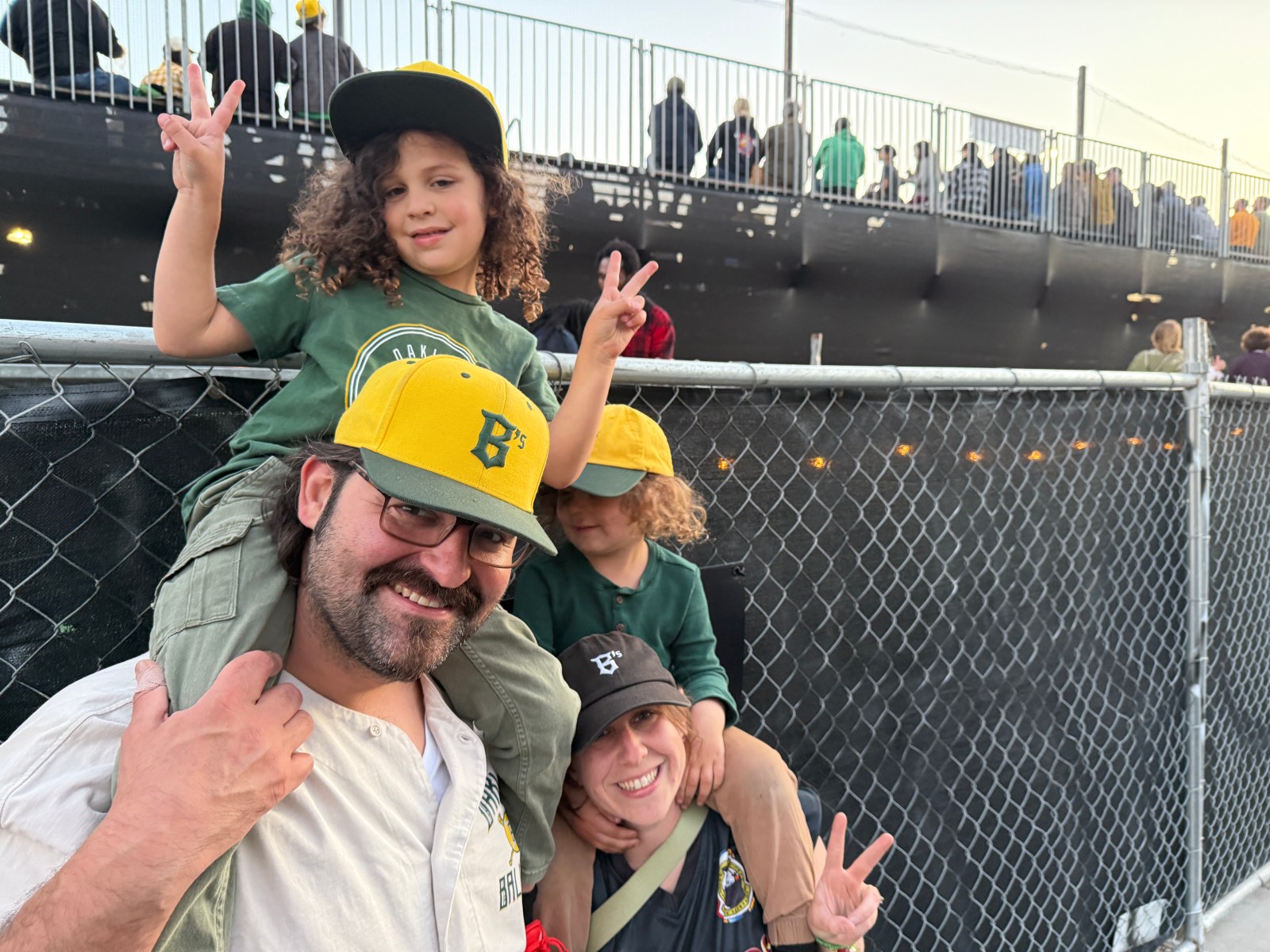 A family at a baseball game, with a father smiling and carrying a child on his shoulders, while another child poses with a peace sign. They are wearing matching green and yellow Oakland Ballers hats and shirts.