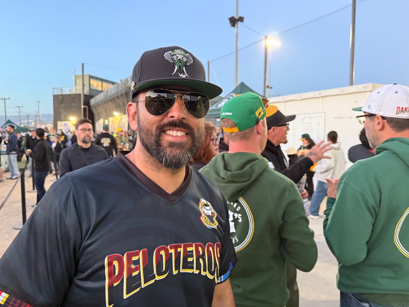 A smiling man wearing sunglasses and a 'Peloteros' jersey stands in a crowd of fans outside a baseball field during an event. The background features a mix of fans wearing various team apparel.