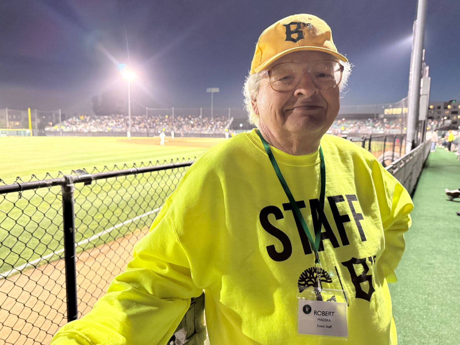 A smiling elderly man wearing a bright yellow 'STAFF' shirt and a matching cap stands by a fence at a baseball field, with a crowd of fans visible in the background.