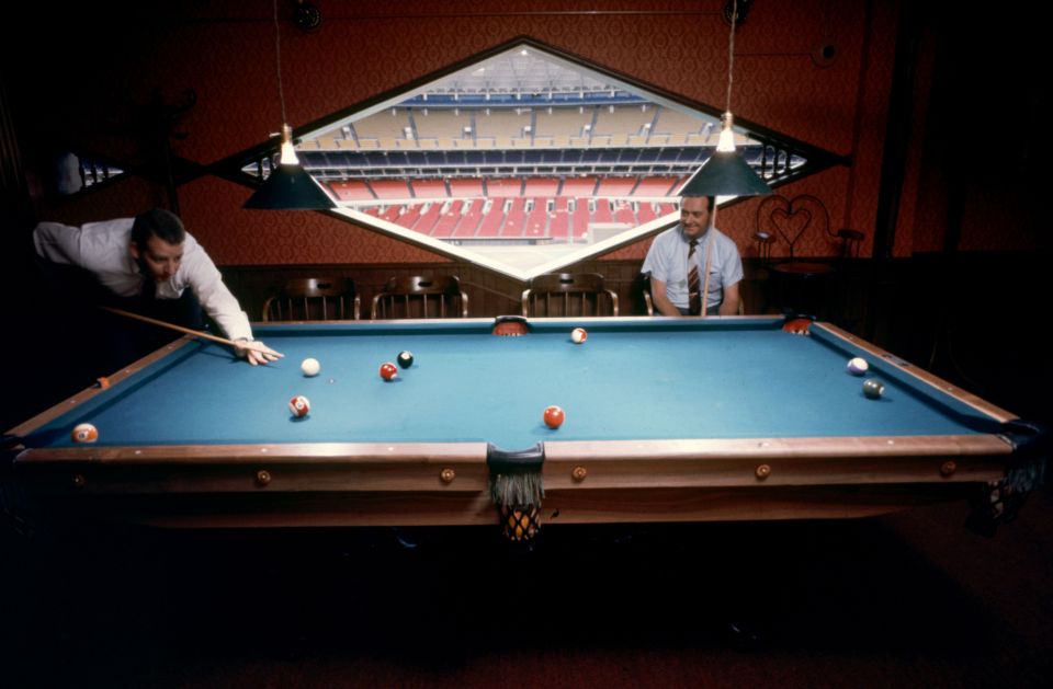 Two men playing billiards in a room overlooking a stadium.