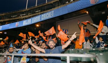 A person stands on stadium stairs, with a cricket field in the background. Some players are on the field, and spectators are in the stands.