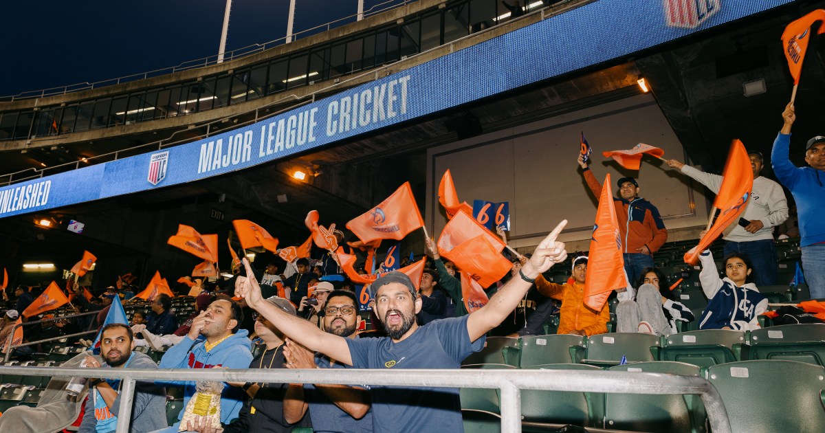A person stands on stadium stairs, with a cricket field in the background. Some players are on the field, and spectators are in the stands.