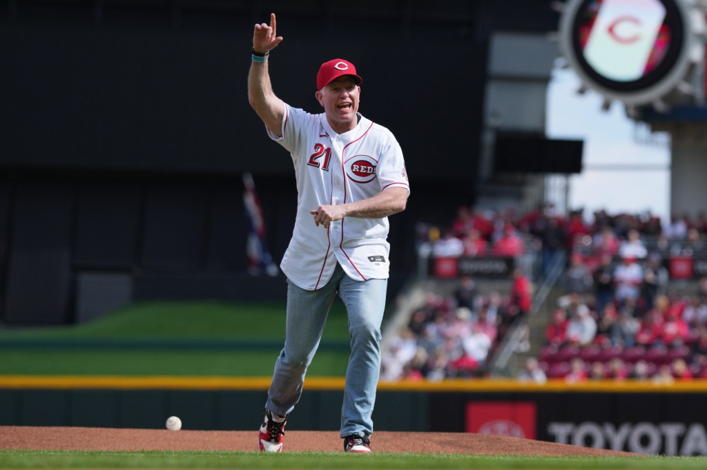 A man in a Cincinnati Reds baseball uniform playfully gestures on the pitcher's mound.