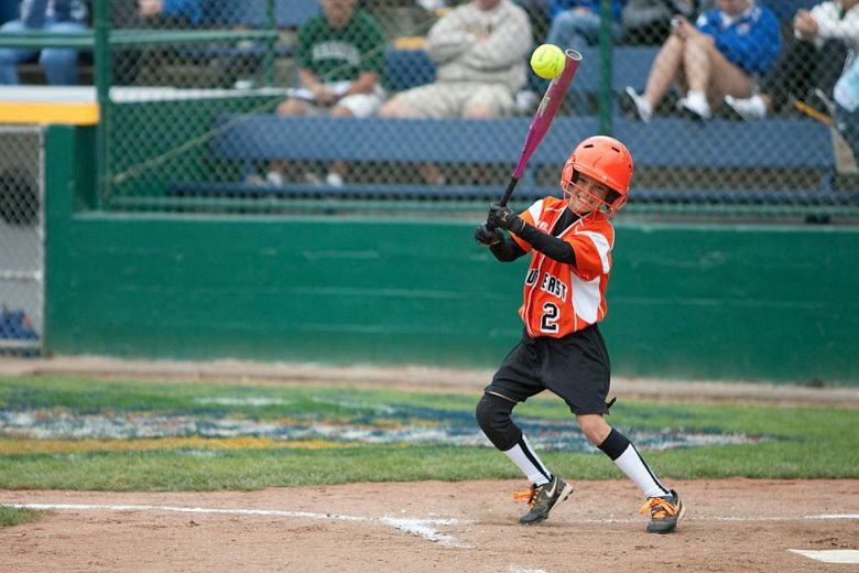 A girl in an orange uniform swings a bat and connects with a yellow softball.