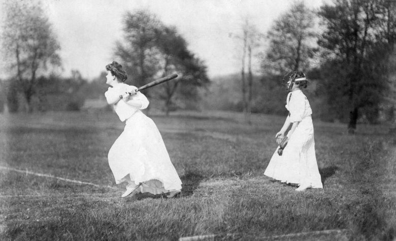 Black and white photo of two women wearing white dresses playing baseball in a grassy field