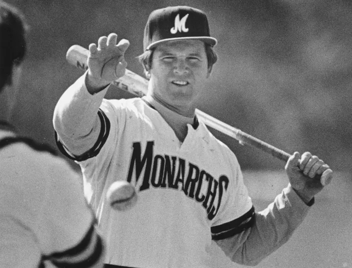 A black-and-white photo of a white man wearing a baseball uniform with the word "Monarchs" on it. He is reaching one hand out to catch a baseball while he holds a baseball bat in the other hand, resting on his shoulder.