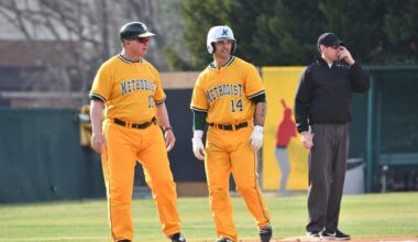 Three white men are standing on a baseball diamond. Two men on the left are wearing yellow Methodist University baseball uniforms. The third man is wearing black.