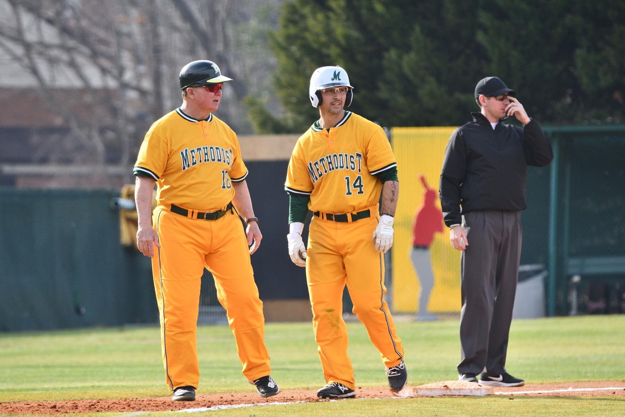 Three white men are standing on a baseball diamond. Two men on the left are wearing yellow Methodist University baseball uniforms. The third man is wearing black.