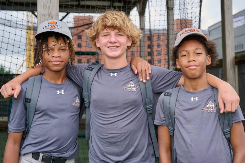 three kids wearing gray shirts and 2 with baseball caps with arms around shoulders smiling for camera