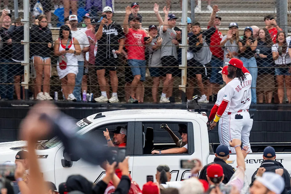 The Cincinnati Reds' Elly De La Cruz waves to fans as he and teammates parade on trucks on the track before the MLB Speedway Classic.