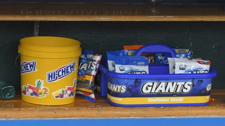 Bags of Giants Sunflower Seeds next to a bucket of Hi-Chew in a baseball dugout.