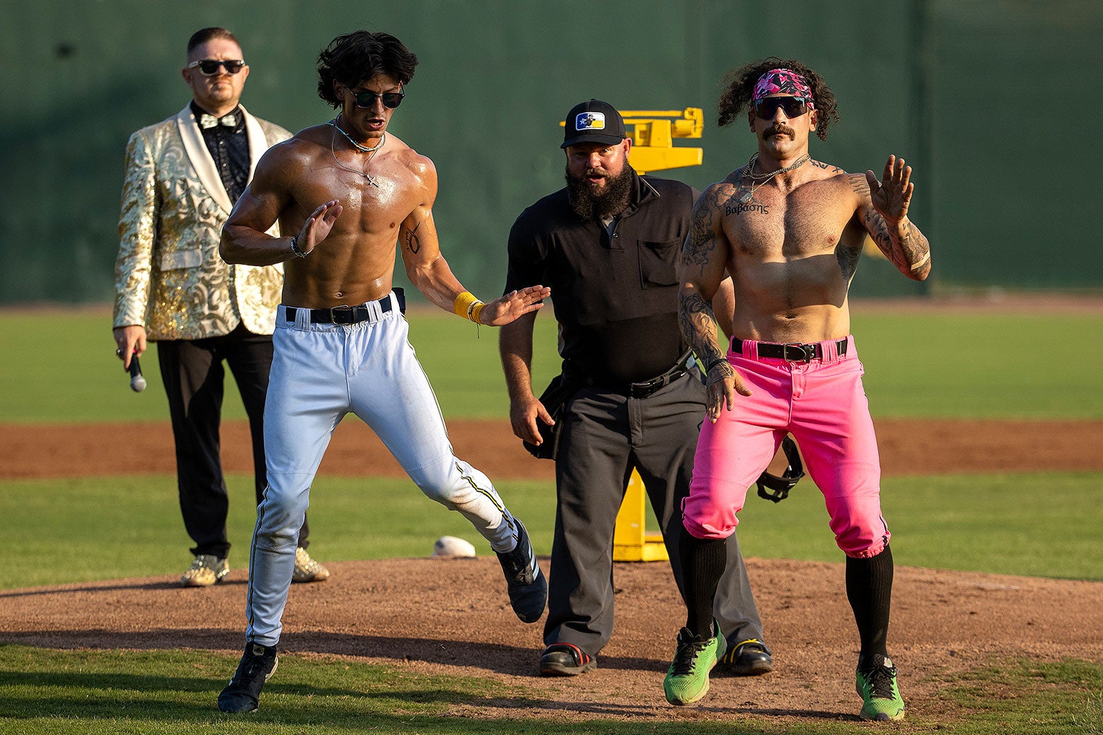 Two shirtless baseball players dance on a field with an umpire egging them on.