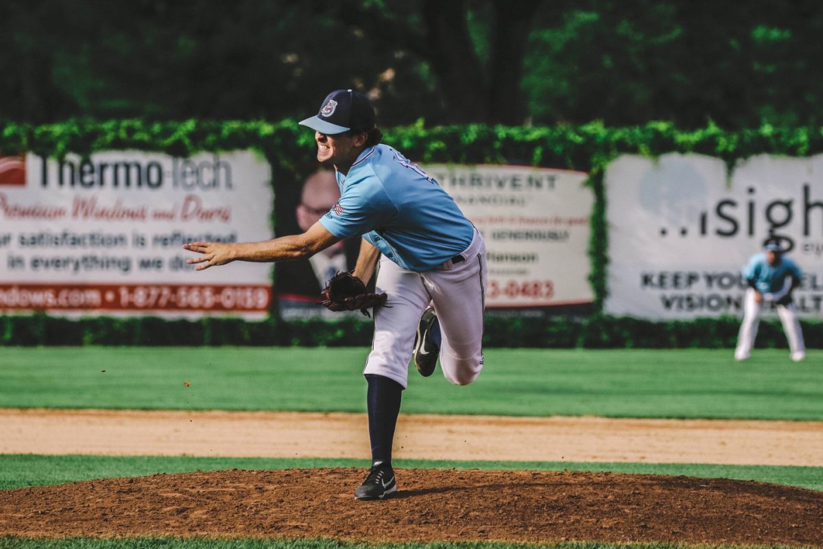 A baseball pitcher in a blue uniform throws a pitch from the mound while another player stands in the outfield. Advertising banners line the fence in the background.