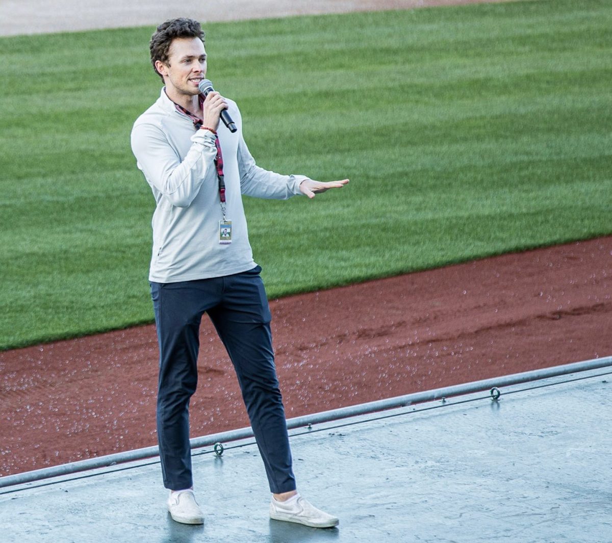 A man stands on a baseball field near the edge of the grass, holding a microphone and speaking, wearing a light jacket, dark pants, and slip-on shoes.