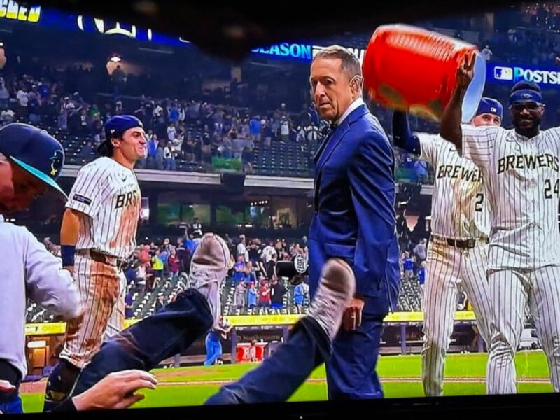 A man in a suit looks surprised as two baseball players in Brewers uniforms dump a cooler of liquid behind him on a stadium field. Other players celebrate, and a person’s legs stick up in the foreground.