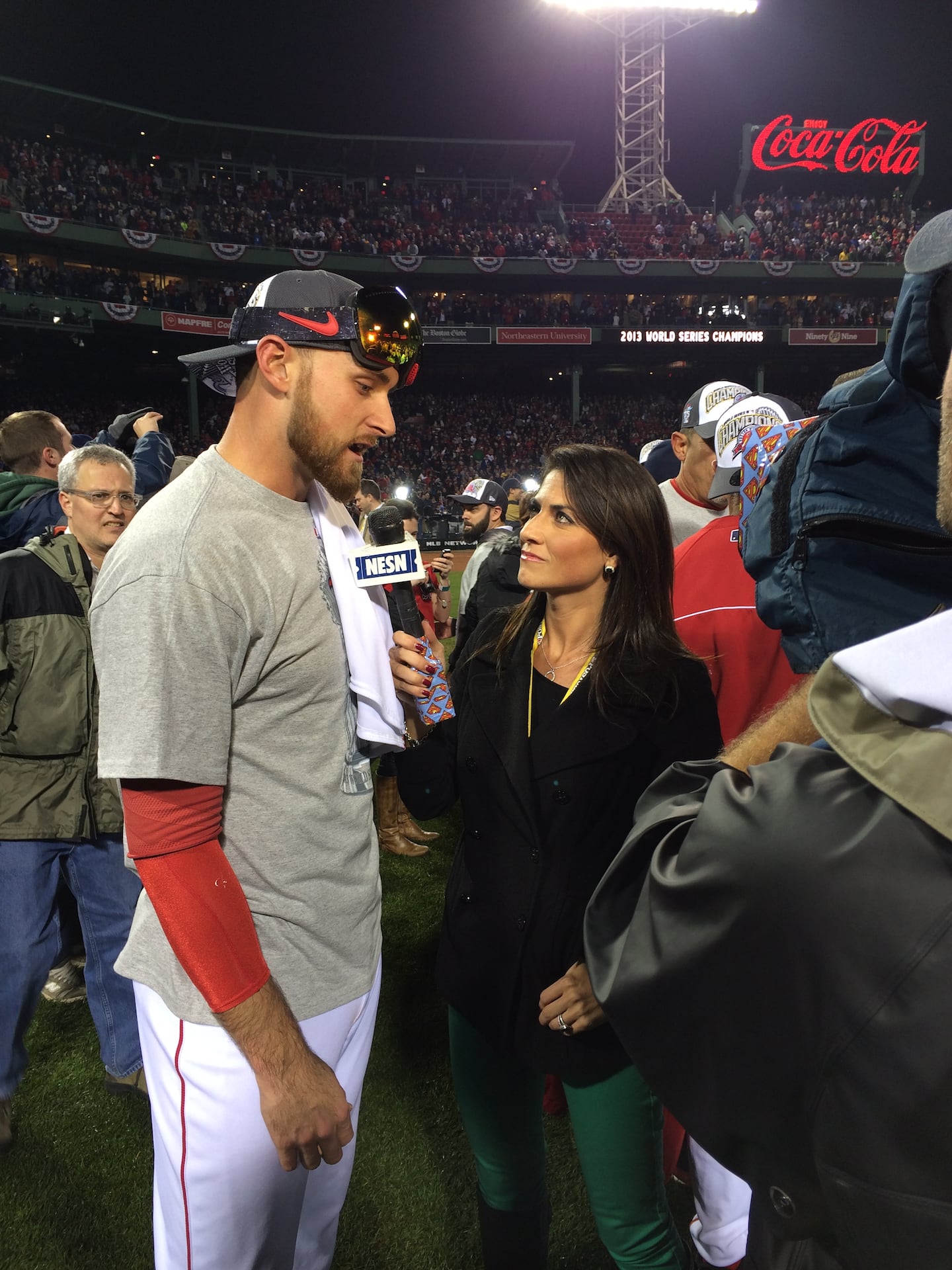 A baseball player stands on the left talking to a female TV reporter holding a microphone. They are on the field surrounded by dozens of people celebrating.