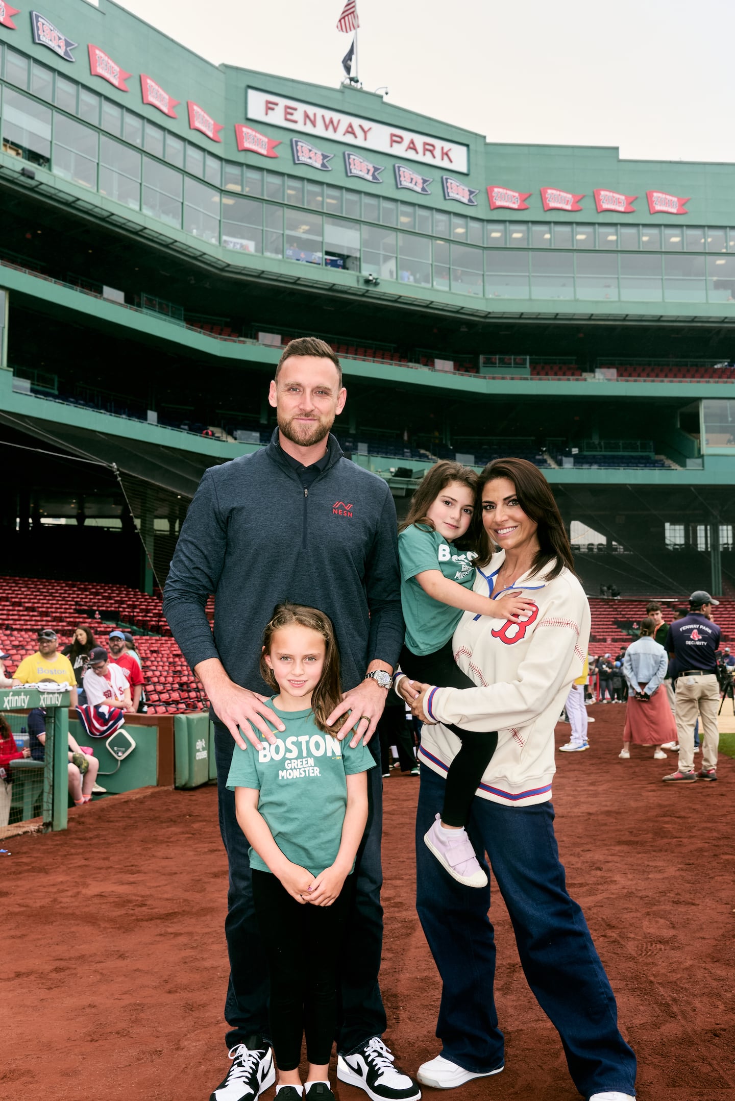 A couple stands with their two young daughters on the dirt on the field at Fenway Park. Red seats behind homeplate are visible in the background.