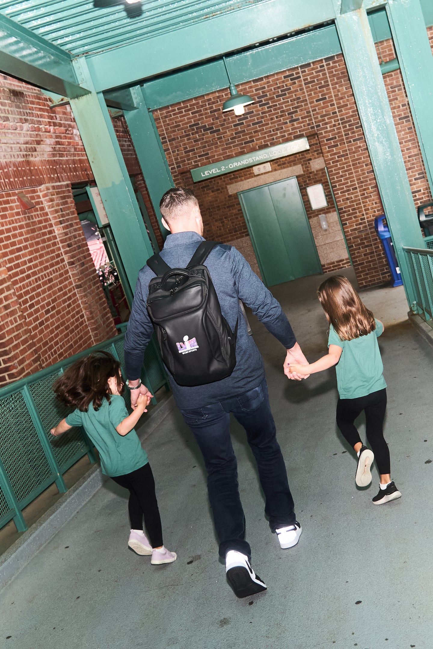 A picture of a man walking holding his daughters' hands as they skip along on either side of him. They are walking down a cement ramp inside Fenway Park.