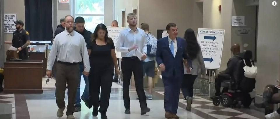 Several people walking in a courthouse, with one man in a blue suit prominently in the foreground.