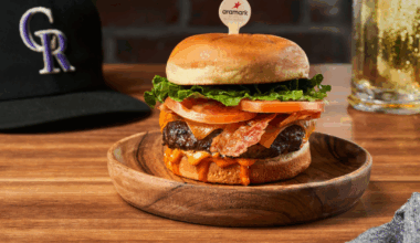 A tall bacon cheeseburger with tomatoes and lettuce is held together by a wooden Aramark skewer on a wooden plate