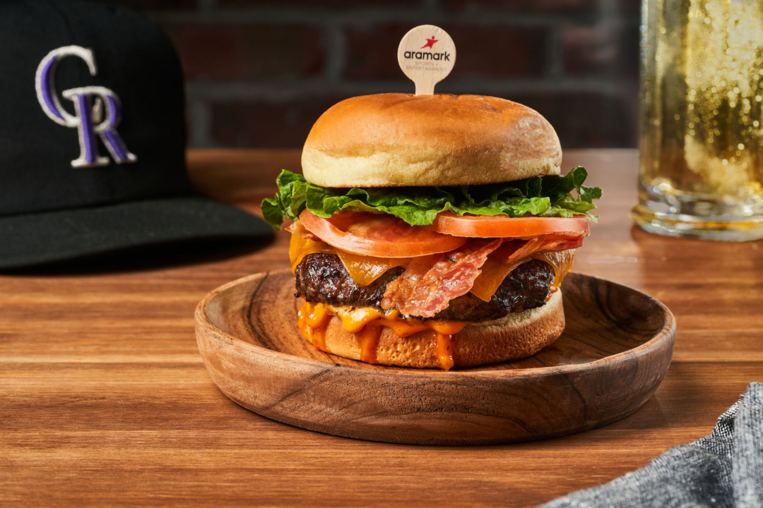 A tall bacon cheeseburger with tomatoes and lettuce is held together by a wooden Aramark skewer on a wooden plate