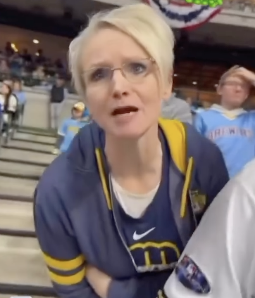 Shannon Kobylarczyk, a woman with short blonde hair and glasses, wearing a blue and yellow hoodie, looks towards the camera with her mouth open as if speaking.