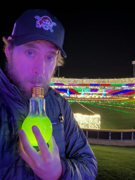 Man holding a green lightbulb cocktail with Christmas decorations from Illumination at Coca Cola Park in the background