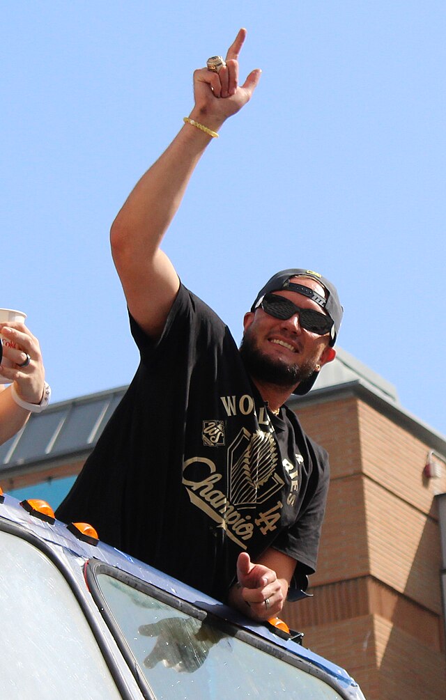 A photo of Dodgers player Miguel Rojas wearing sunglasses, a black backwards baseball cap, and his black 2025 World Series Champion t-shirt, smiling and holding his hand up and pointing upwards as he leans out of a car at the victory celebration parade.
