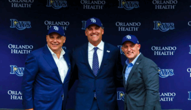 Three members of the Tampa Bay Rays’ new ownership group pose together wearing team hats at a press conference backdrop featuring the Rays and Orlando Health logos.