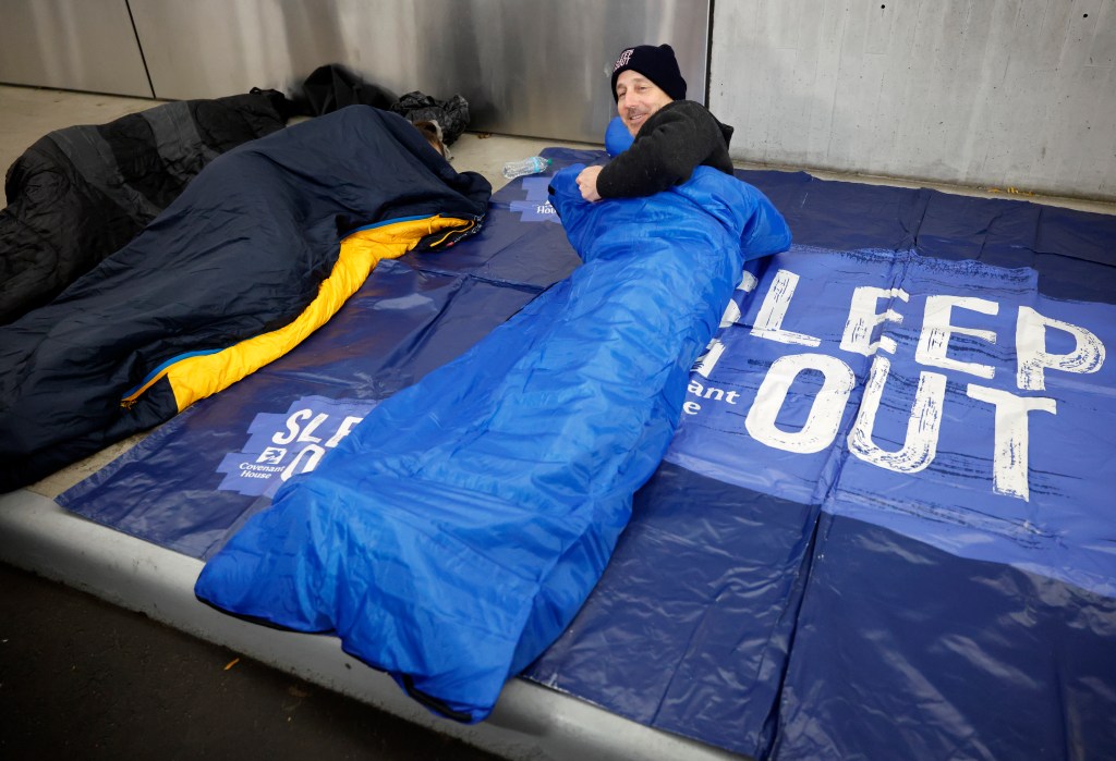 Yankees GM Brian Cashman in a blue sleeping bag on a blue mat that says "Sleep Out" for the Covenant House Sleep Out event.