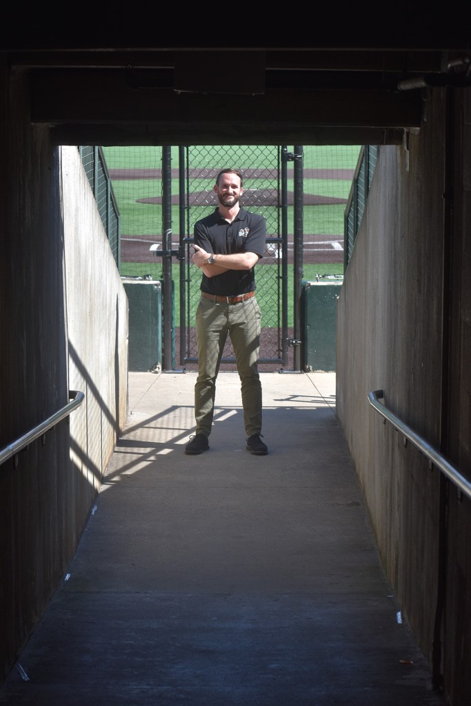 Night Train stands in one the halls leading out to the baseball stadium. He stands in the sun with his arms crossed, while the hallway is in shadows. The field is partially visible behind him. He wears green pants and a black polo shirt.