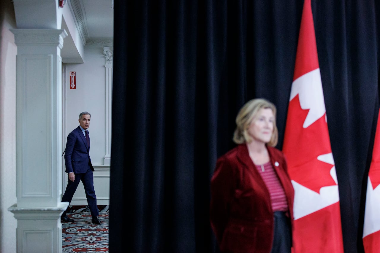 A man in a suit and tie enters a hotel ballroom, with a woman standing in front of a dark curtain near Canadian flags.