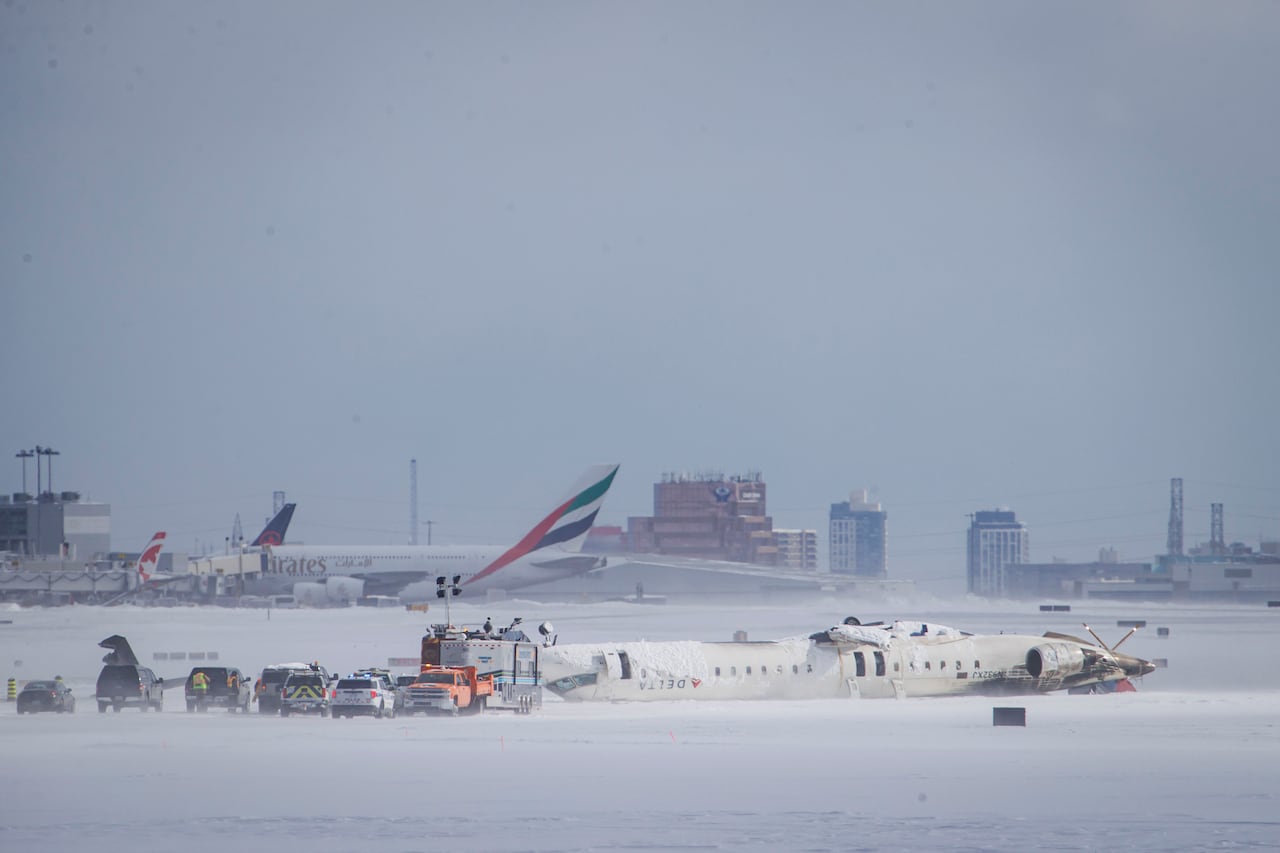 A plane lying upside down on a snowy tarmac, with vehicles and crews nearby.