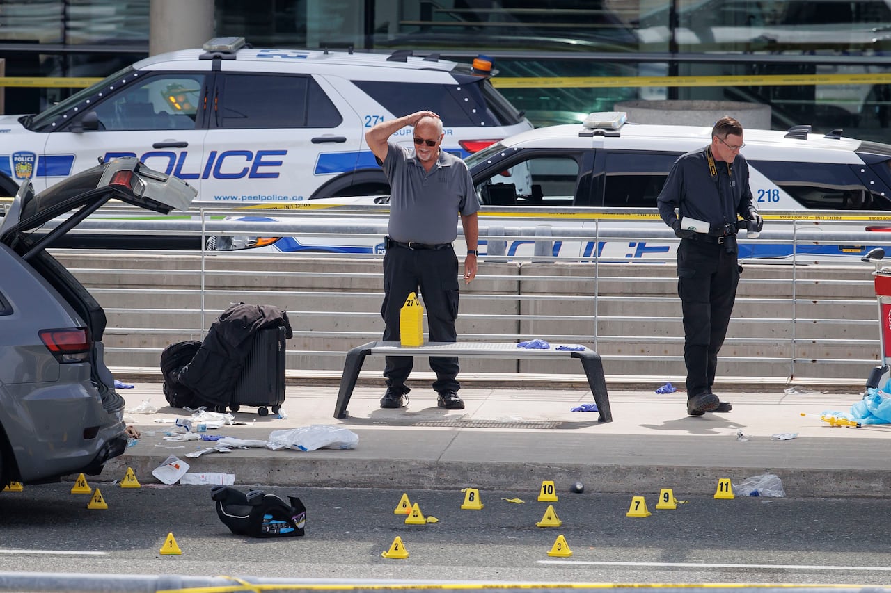 Two investigators including one raising his hand to his head looking perplexed outside an airport and evidence markers naer an SUV.