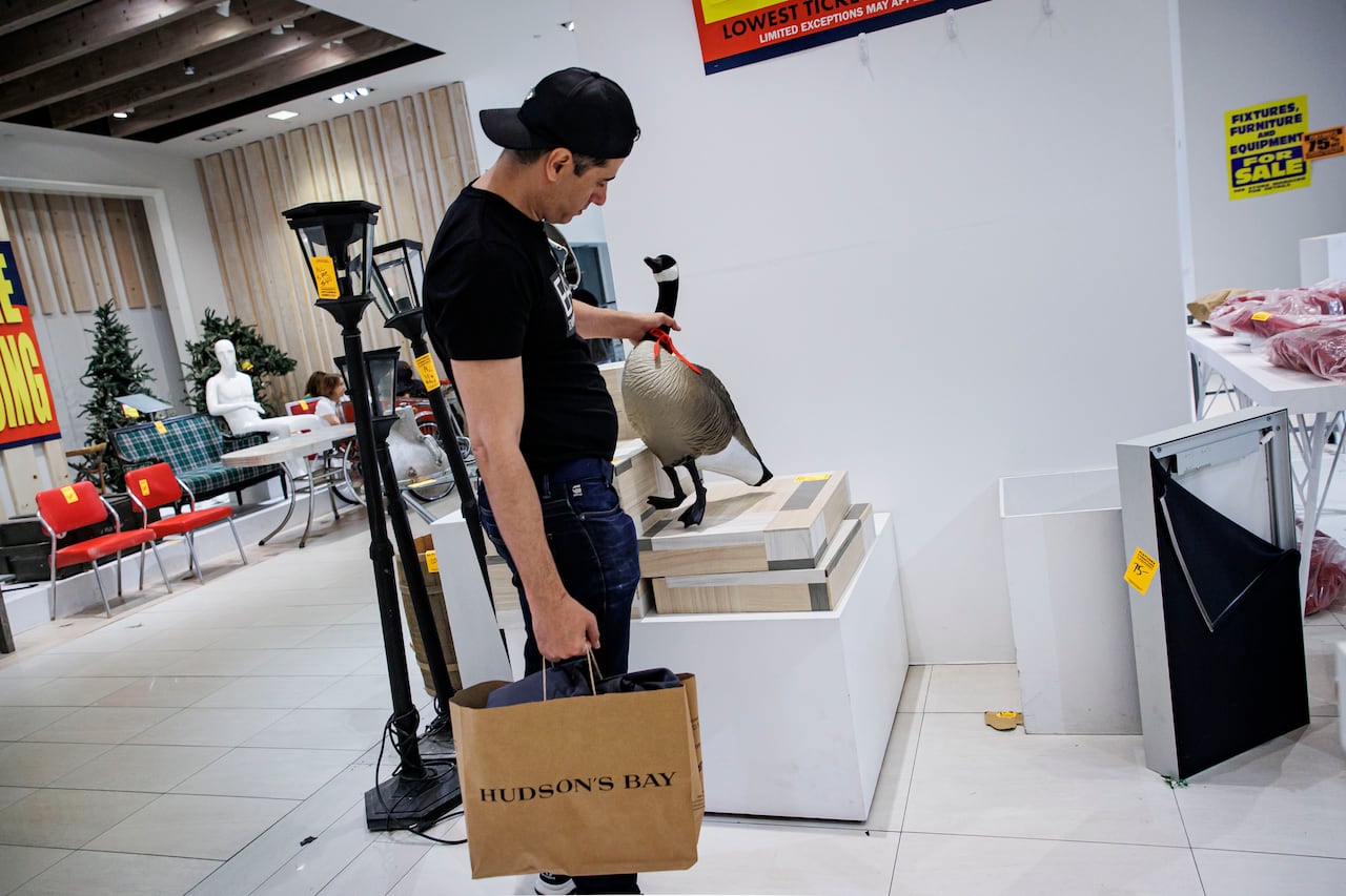 Male shopper holding paper bag that reads Hudson's Bay holding with his other hand a Canada geese for sale in a store surrounded by other sale items.
