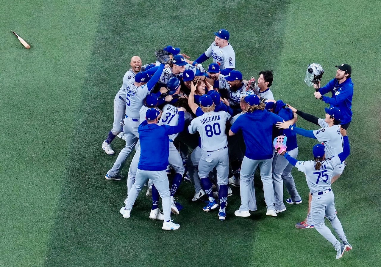 Baseball team rush to one another on the mound, celebrating their pitcher, with a broken bat near them.