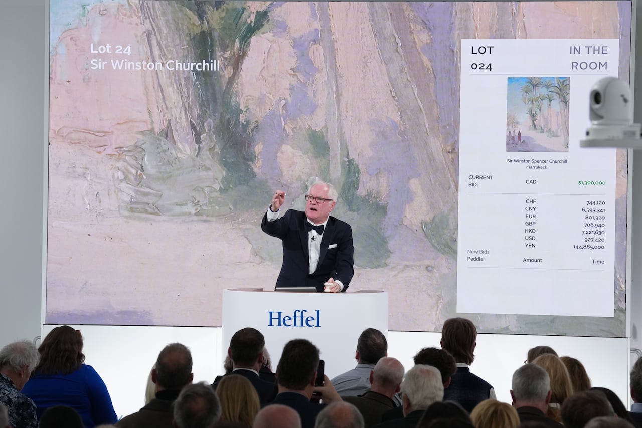 A man in a tuxedo presiding over an auction with a painting near him on a screen and bidders in the foreground.