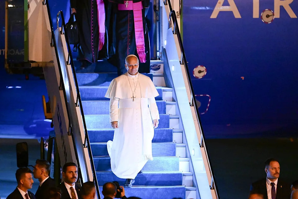 A religious leader in white papal attire descends an airplane staircase, surrounded by officials and media at a public event