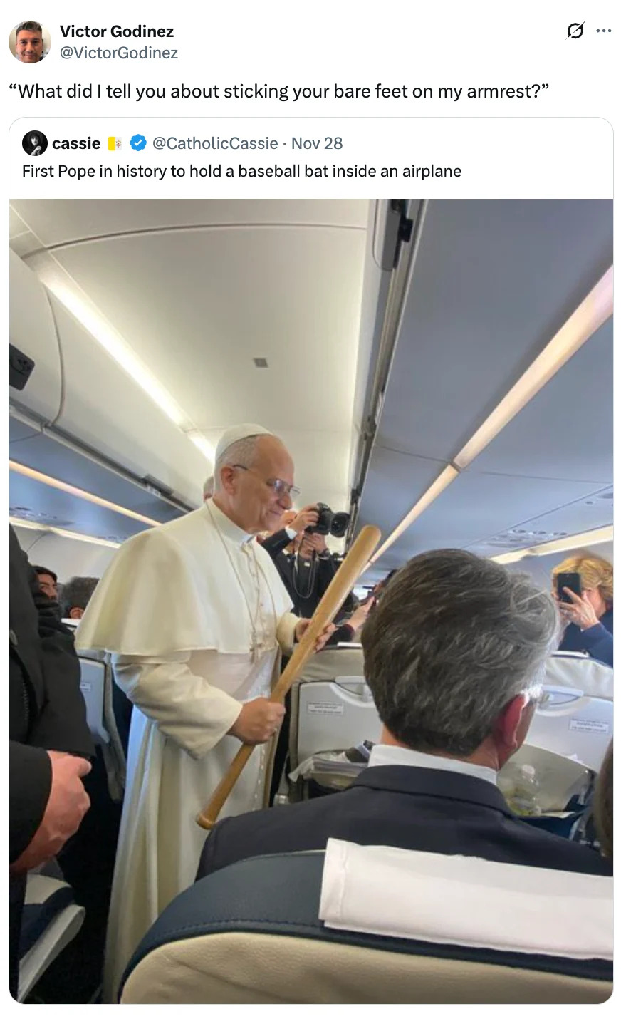 Pope holds baseball bat on airplane, surrounded by people, including a seated person in a suit