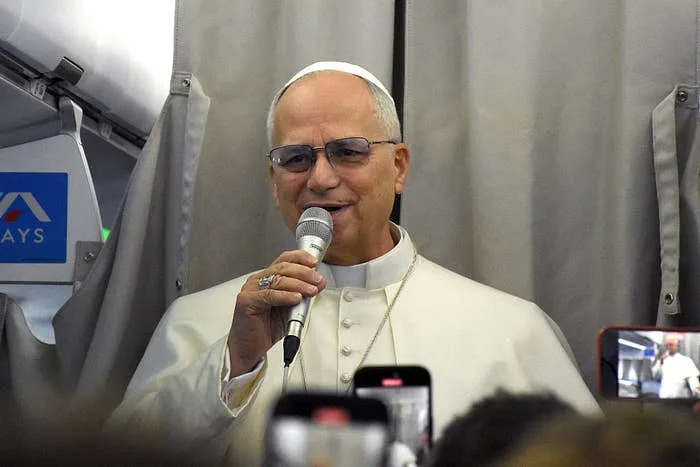 A man dressed in Papal attire speaks into a microphone while on an airplane, surrounded by people holding smartphones