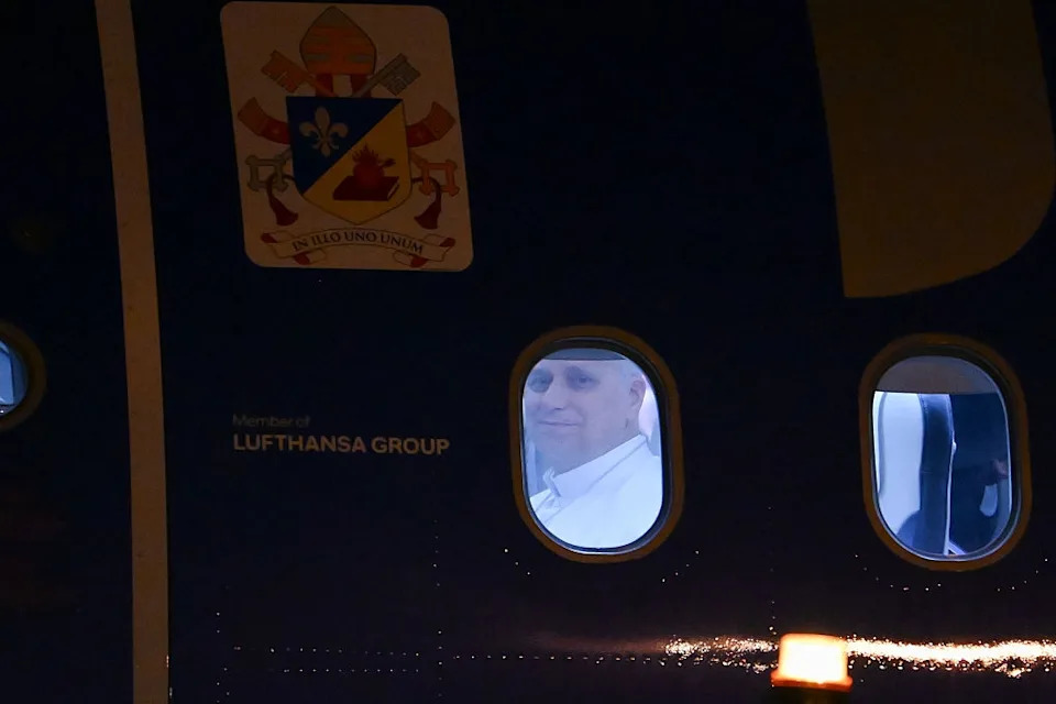 Person smiling slightly, looking out from an airplane window. The plane features a prominent emblem and "Lufthansa Group" text visible