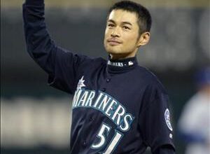 A young asian man in a blue baseball uniform waves his batting helmet in the air. His jersey says mariners above the number fifty one.