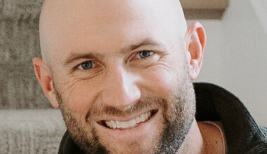 A smiling adult man with a bald head and trimmed beard sits indoors on carpeted stairs, wearing a dark zip-up sweater. He is looking directly at the camera.”