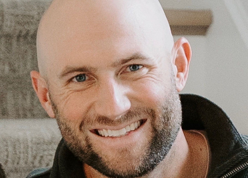 A smiling adult man with a bald head and trimmed beard sits indoors on carpeted stairs, wearing a dark zip-up sweater. He is looking directly at the camera.”